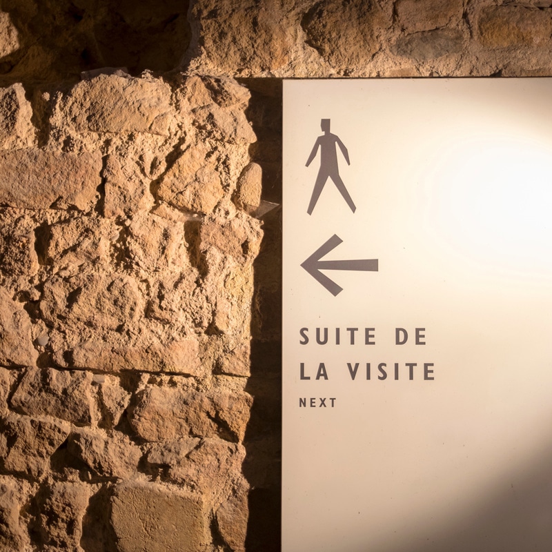 Signalétique Musée : Panneau Suite de la Visite et Next Panneau moderne sur mur de pierres avec texte SUITE DE LA VISITE NEXT, flèche et pictogramme de marcheur.